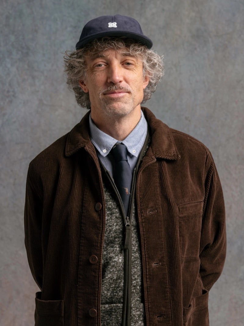 Man in a brown corduroy jacket over a shirt and tie, wearing a navy cap, for Kestin Fall 2026
