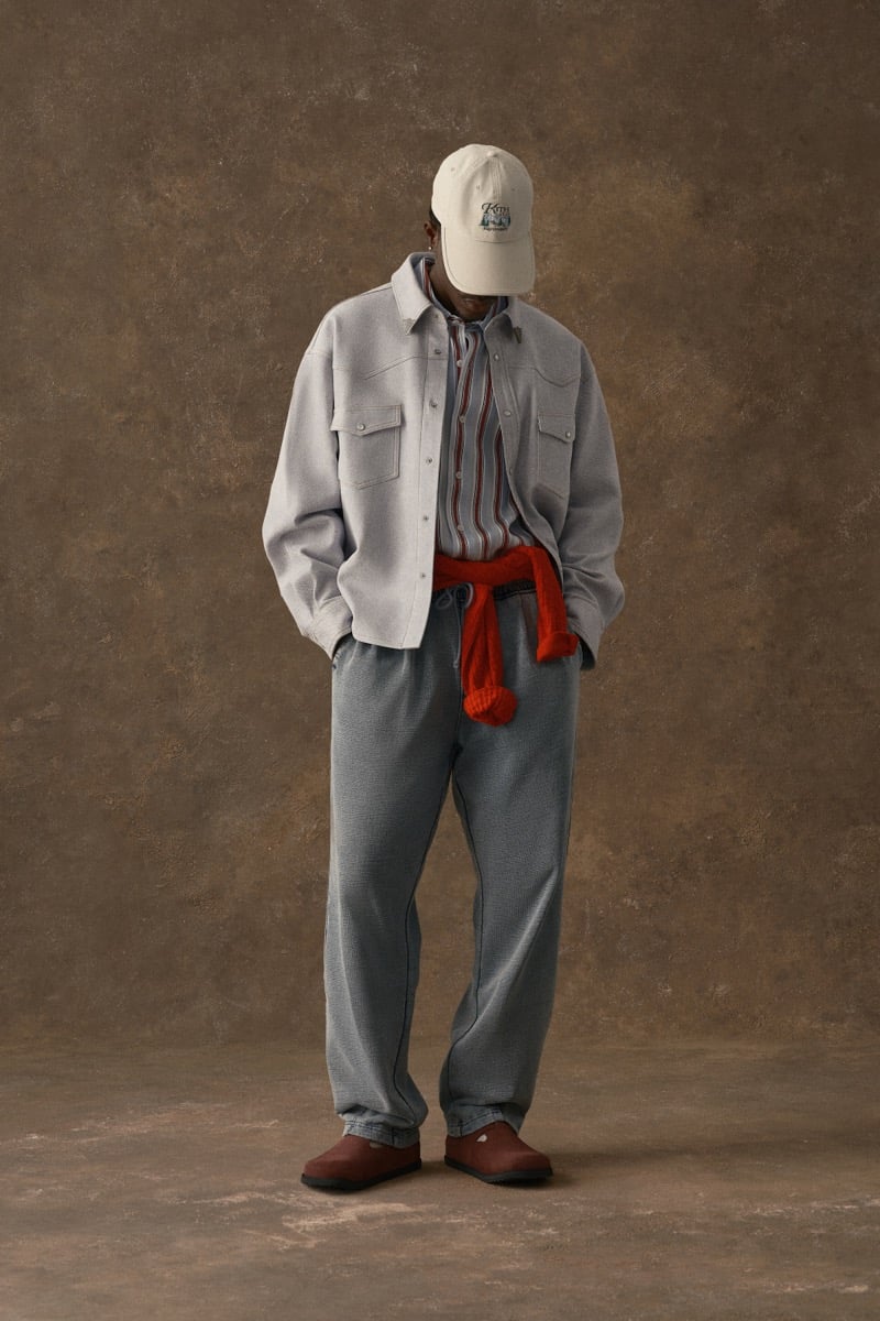 Man in a gray jacket with a red scarf tied at the waist, wearing a cream cap, for Kith Fall 2026