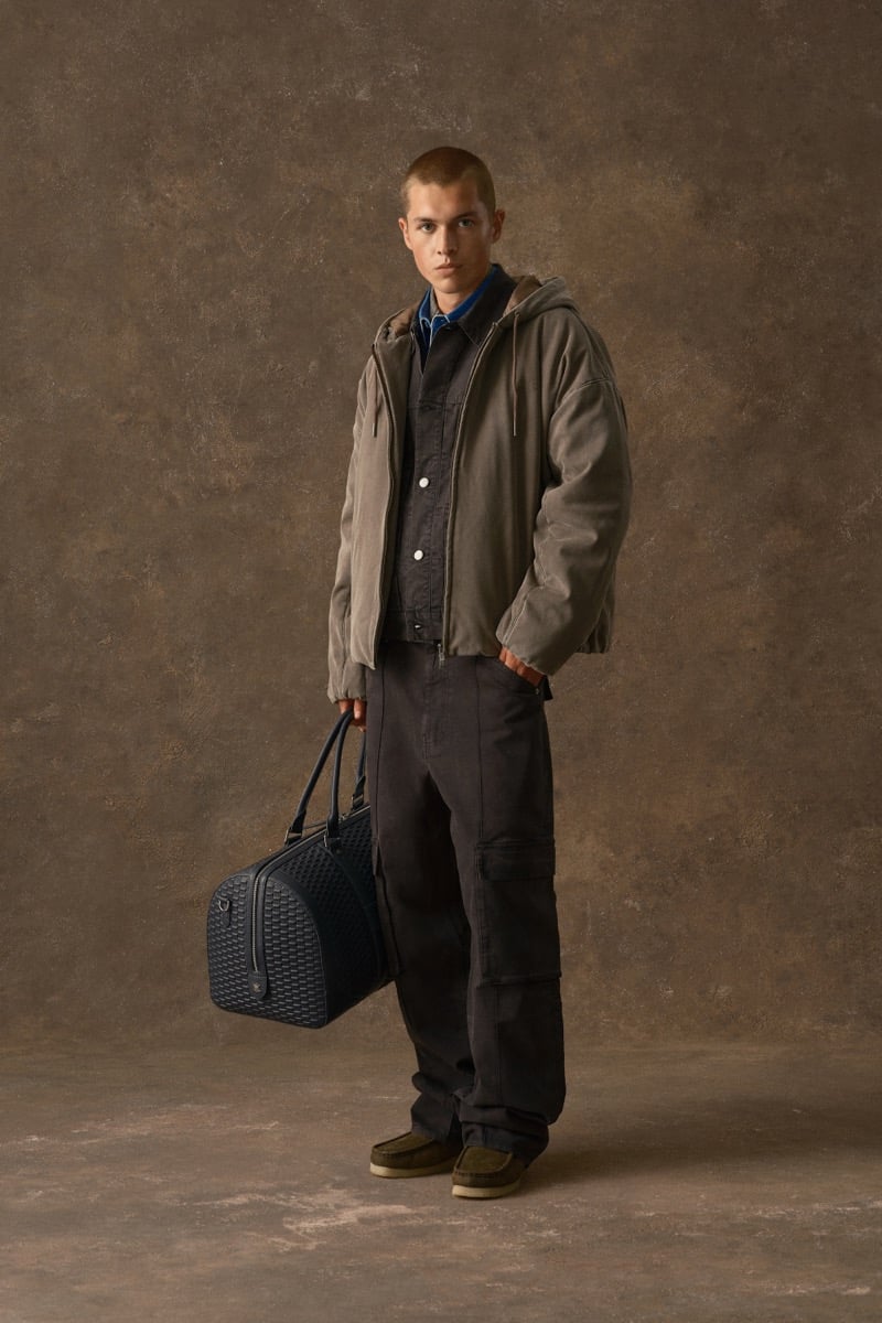 Man in an olive hooded jacket over a blue collared shirt, carrying a woven duffel bag, for Kith Fall 2026