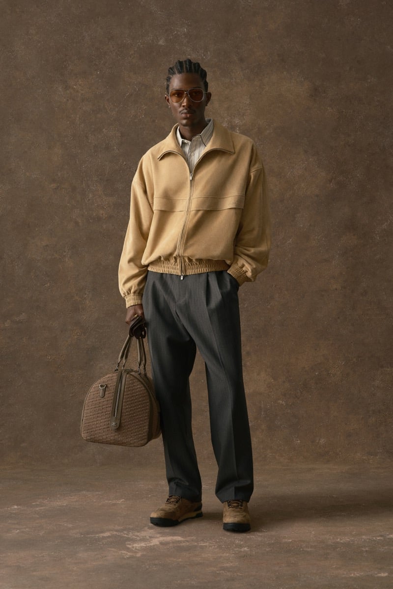 Man in a tan bomber jacket over a striped shirt, carrying a woven duffel bag, for Kith Fall 2026