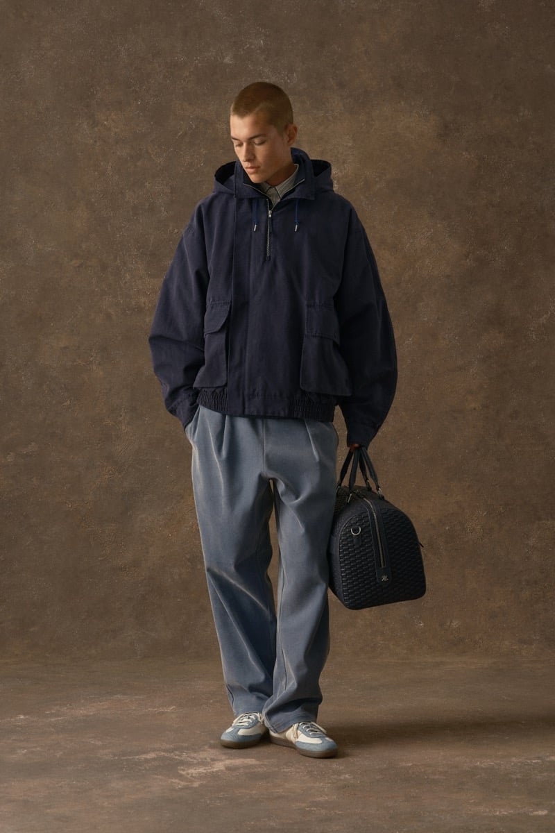 Man in a navy hooded jacket, carrying a bag over blue-gray trousers, for Kith Fall 2026