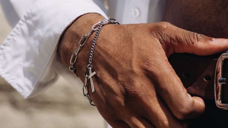 A man's wrist wearing a silver chain bracelet with a cross charm, hand resting on a belt