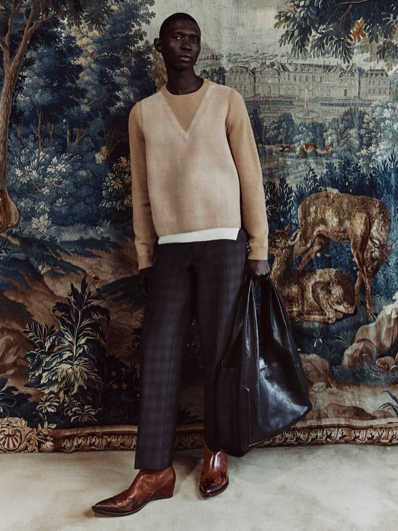 Fernando Cabral in a tan Maison Margiela sweater and plaid trousers, holding a leather tote in front of a tapestry backdrop, wearing brown western boots with flame detailing