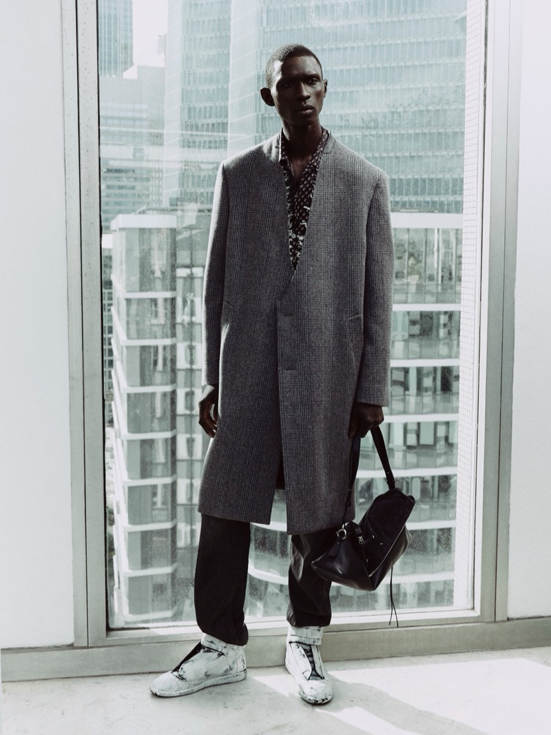 Fernando Cabral in a gray checked Maison Margiela overcoat over a printed shirt, dark trousers, and paint-splattered sneakers in front of floor-to-ceiling windows