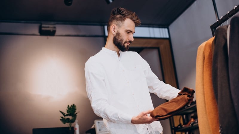 A bearded man in a white shirt examines a brown jacket in a menswear shop, a rack of clothing beside him