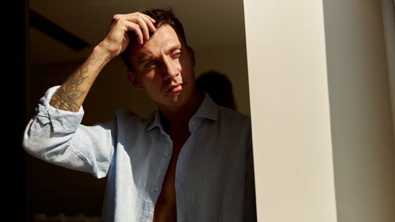 A man in an unbuttoned linen shirt runs a hand through his hair in a sunlit doorway, a tattoo visible on his forearm