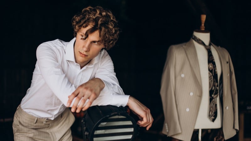 A curly-haired man in a white shirt leans against a case in a dim studio, a pinstripe suit and patterned tie displayed on a mannequin behind him