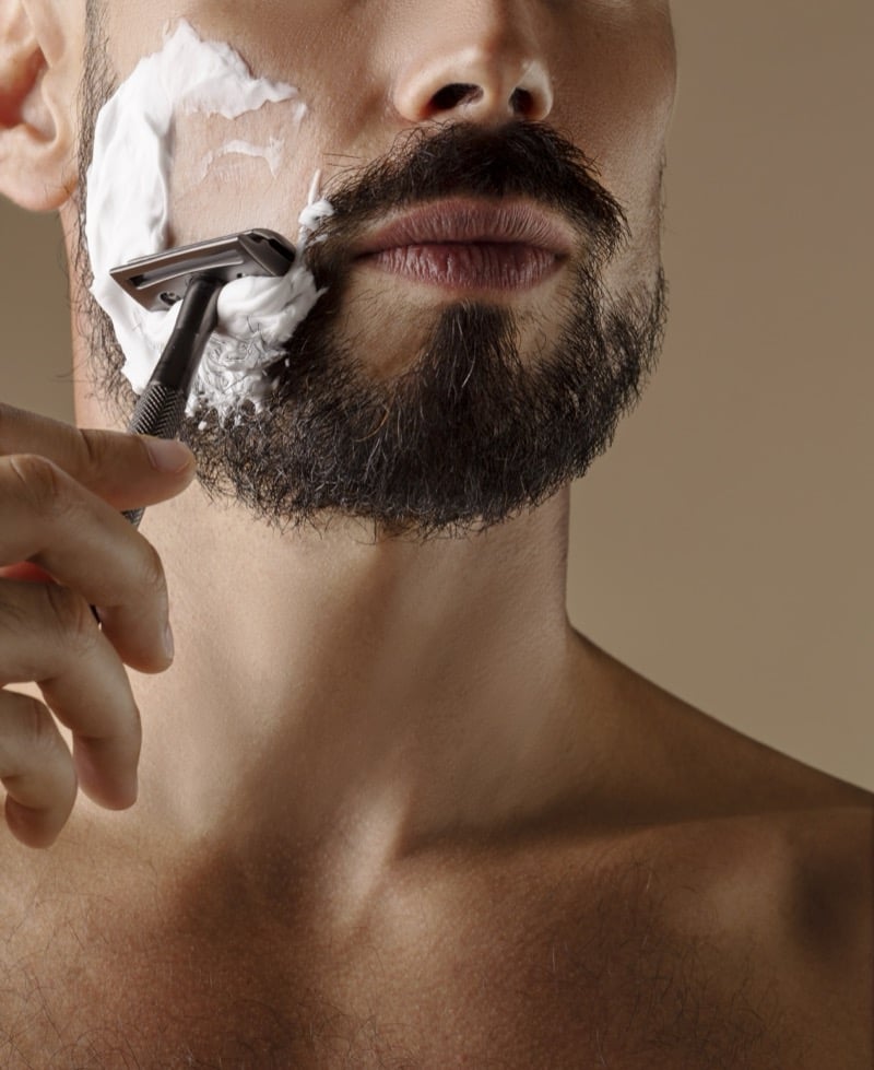 Bare-chested man shaving the edge of his dark beard with a safety razor and a patch of shaving cream