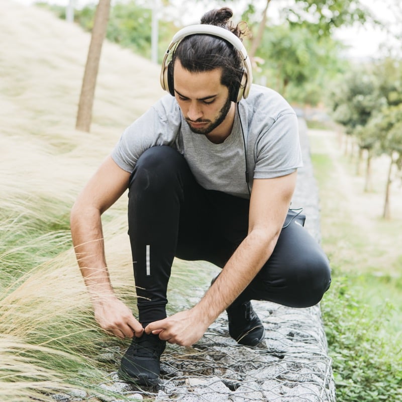 A man wearing headphones crouches to tie his running shoes on a path lined with tall grass