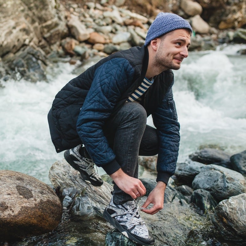 A man in a beanie and quilted jacket crouches on rocks beside a rushing river, tying his shoes