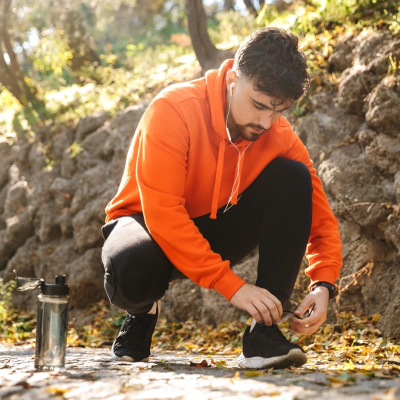 A man in an orange hoodie crouches on a leaf-covered path to tie his black running shoes