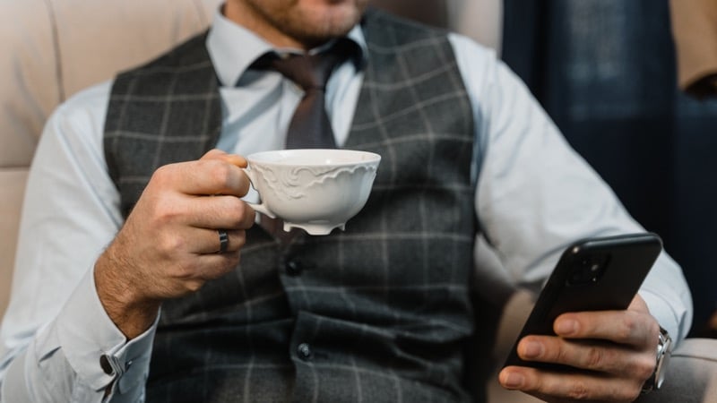 A man wearing a wedding band holds a teacup in one hand and a phone in the other