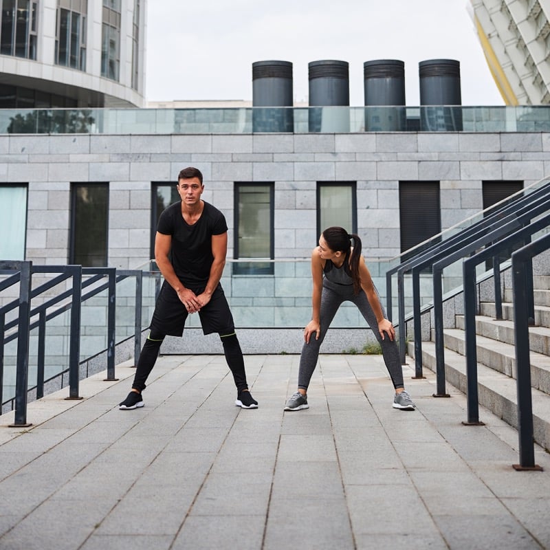 A man and woman stretch on an outdoor staircase between glass and stone buildings