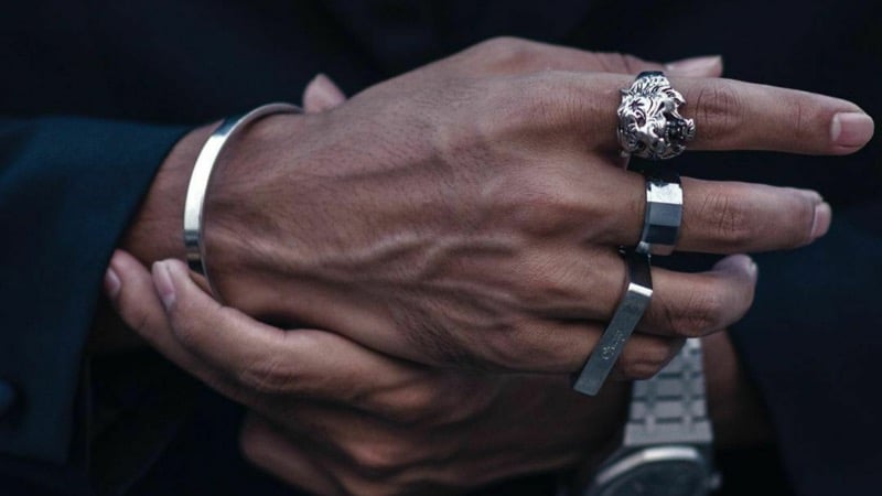 A man's clasped hands wearing a silver cuff bracelet, rings, and a watch against a dark suit
