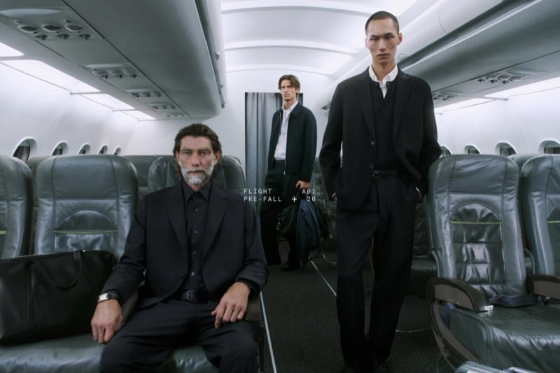 Patrick Petitjean sits with a weekender bag as Justin Eric Martin and Siquan Wang stand behind him in black tailoring inside an airplane cabin