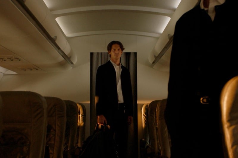 Justin Eric Martin stands in the aisle in a dark jacket and white shirt, holding a bag, seen past rows of seats