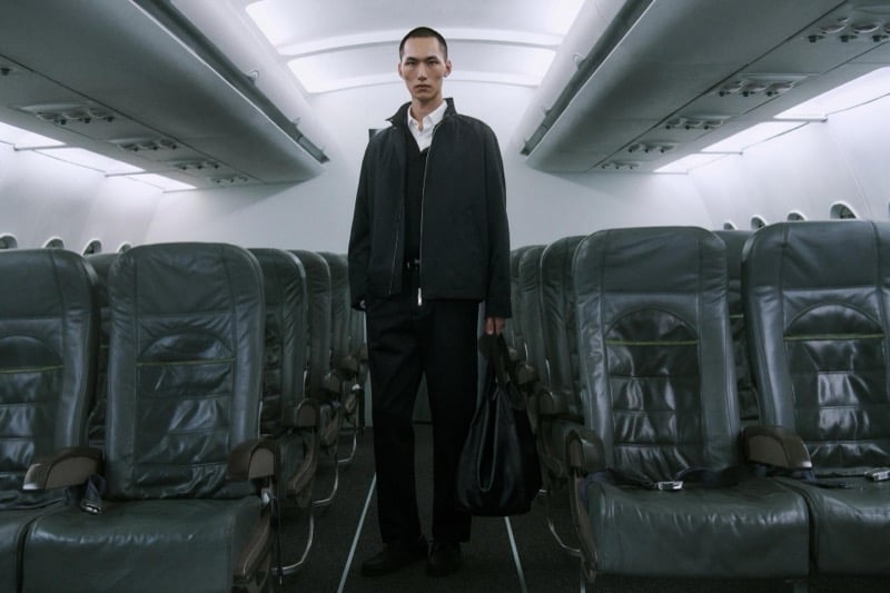 Siquan Wang stands in the aisle in a black zip-front jacket, holding a large tote bag