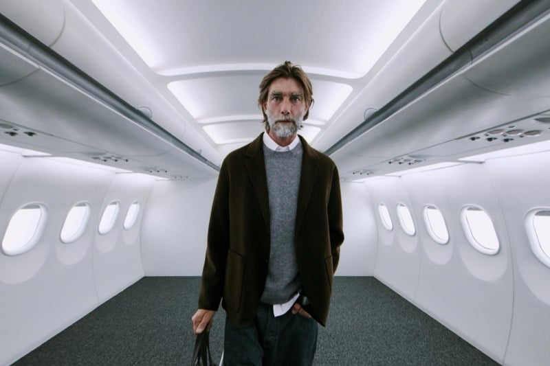 Patrick Petitjean stands alone in the aisle in a brown jacket over a gray sweater, holding a small bag