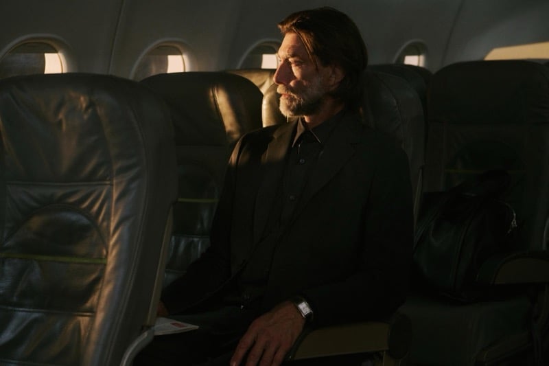 Patrick Petitjean sits alone in a dark suit gazing out an airplane window lit by late afternoon light
