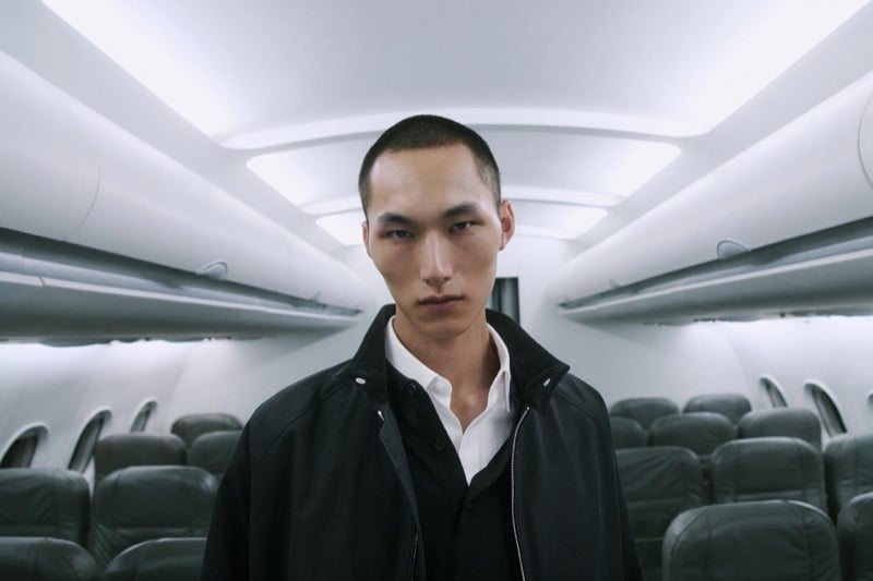 Siquan Wang looks directly at the camera in a black zip-front jacket and white shirt inside the airplane cabin