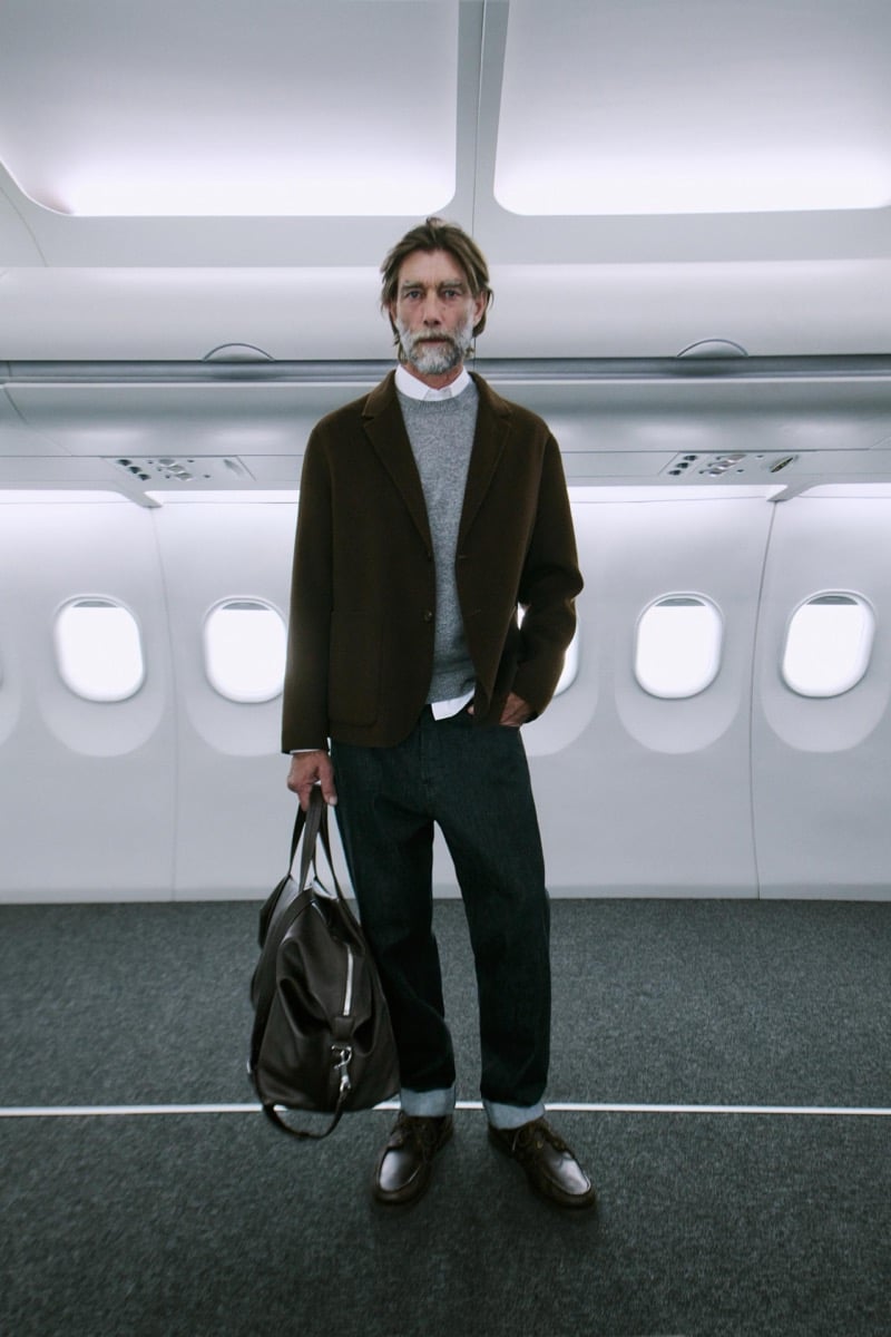 Patrick Petitjean stands in a brown jacket, gray sweater, and cuffed jeans, holding a weekender bag