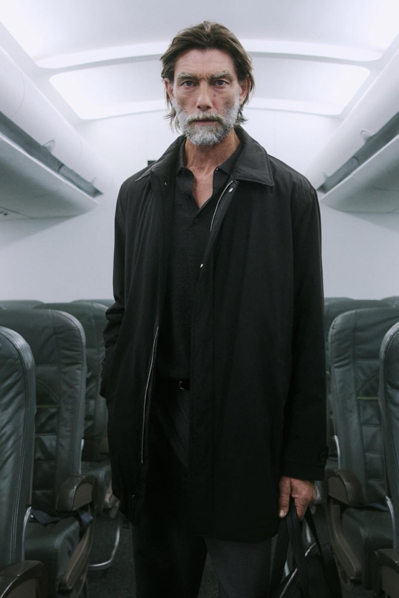 Patrick Petitjean stands alone in a black zip-front coat and black trousers, holding a bag, inside the airplane cabin