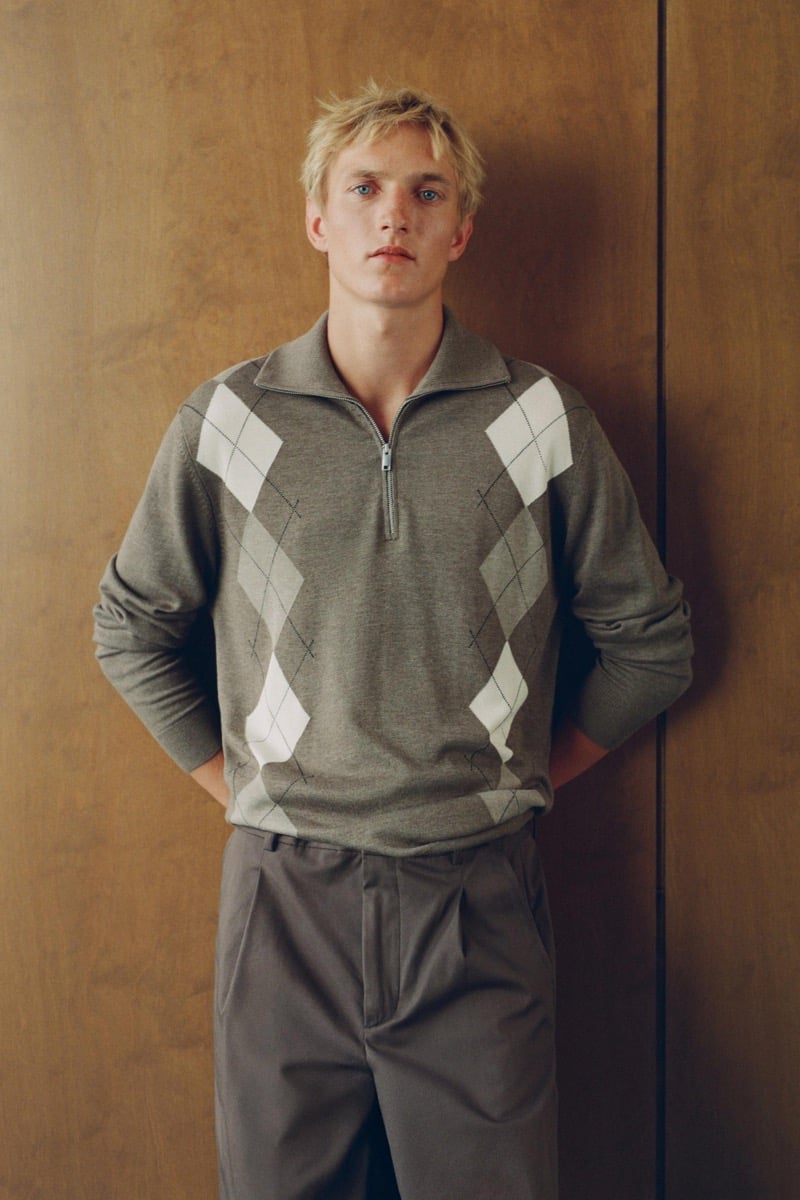 Pierrick Grégoire stands with hands clasped behind his back in a gray argyle half-zip sweater and pleated trousers