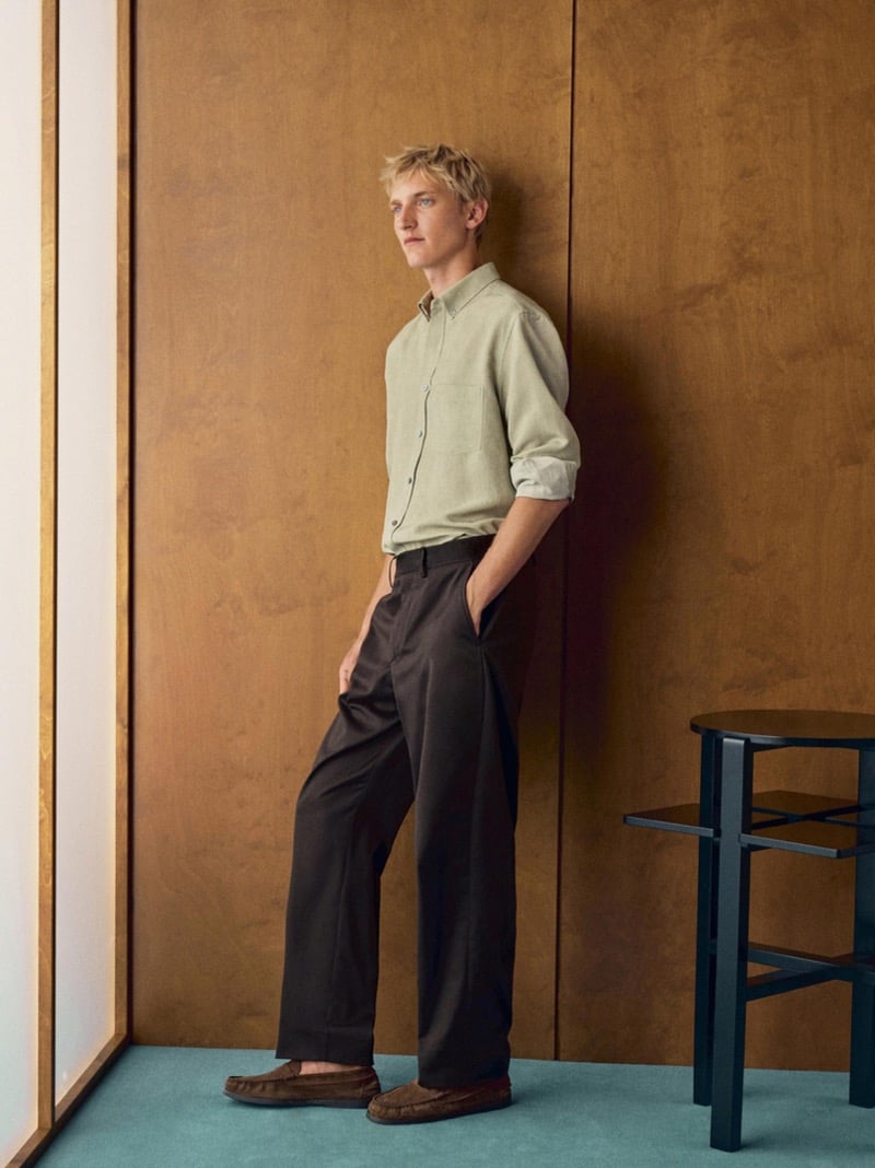 Pierrick Grégoire leans against a wood panel wall in a sage shirt, dark brown pleated trousers, and suede loafers