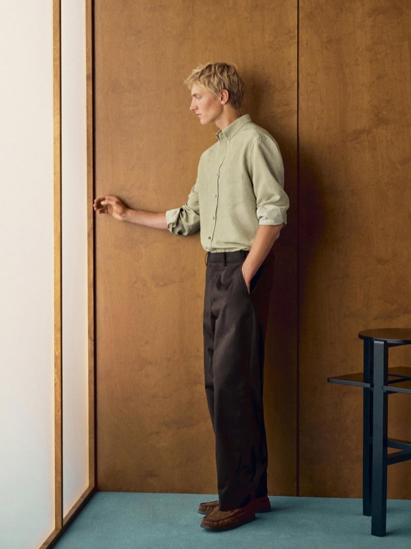 Pierrick Grégoire reaches toward a door in profile, wearing a sage shirt, dark brown trousers, and suede loafers