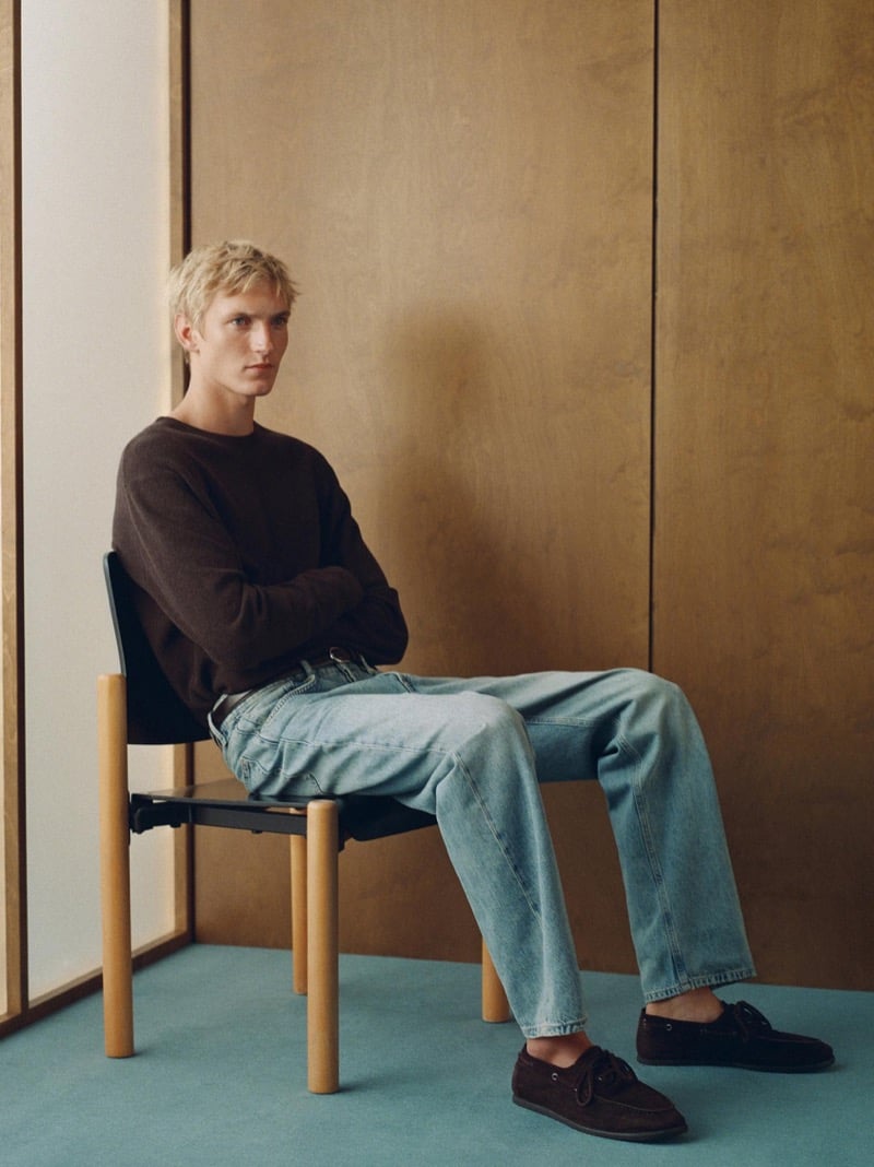 Pierrick Grégoire sits with arms crossed in a dark brown crewneck sweater, light wash jeans, and suede boat shoes