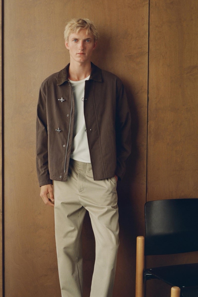 Pierrick Grégoire wears an open brown workwear jacket with cross-stamped hardware over a white t-shirt and stone trousers