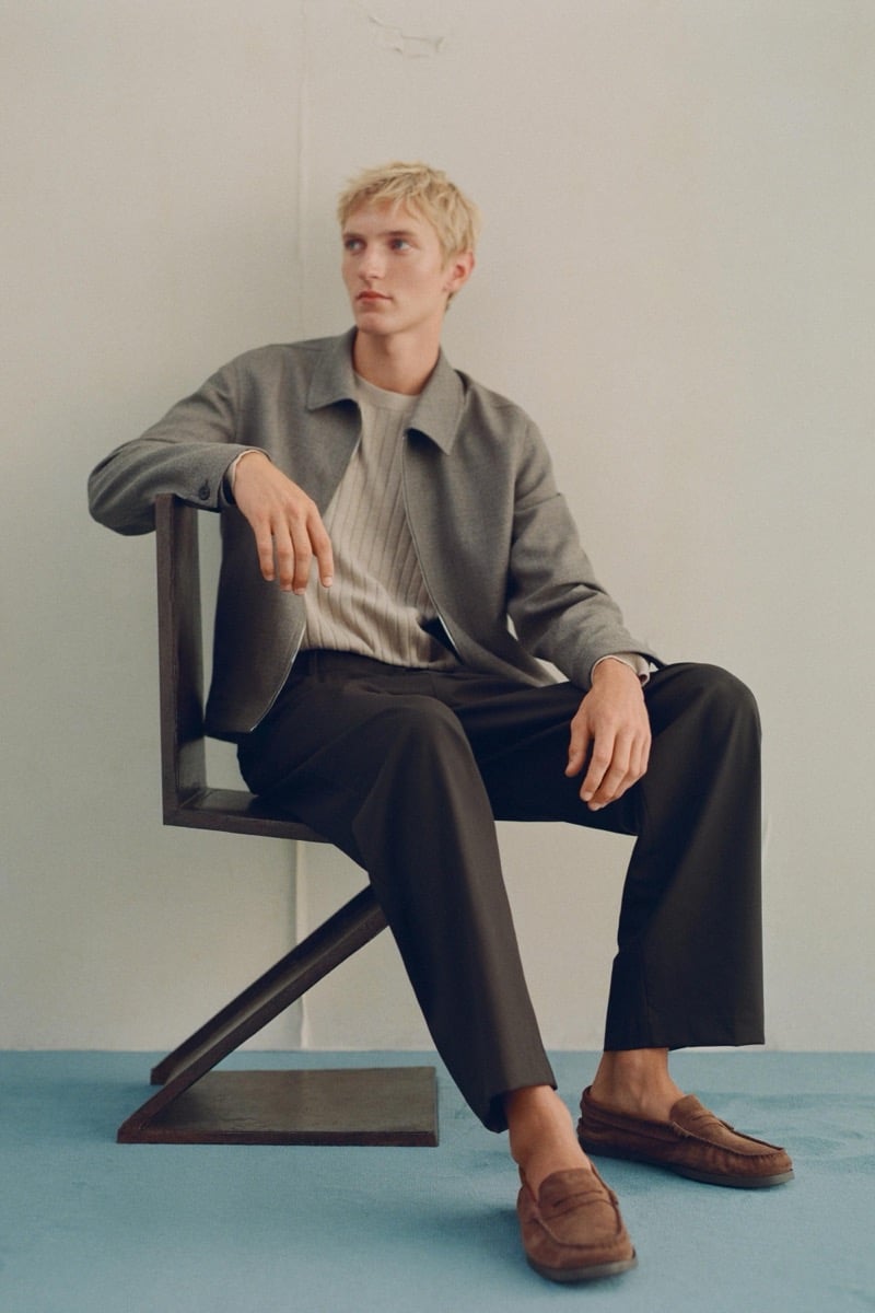 Pierrick Grégoire sits in an angular wood chair wearing a gray cropped jacket over a ribbed knit sweater with dark trousers and suede loafers
