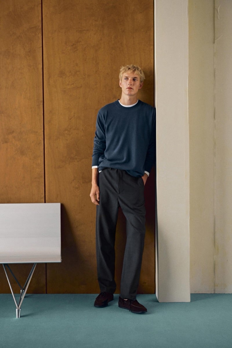 Pierrick Grégoire leans against a wall in a blue crewneck sweater with dark trousers and burgundy suede shoes