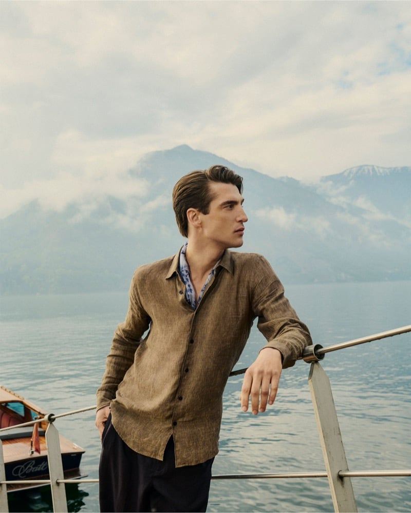 Anatol Modzelewski leans on a boat railing on Lake Como wearing a brown linen shirt over a printed shirt