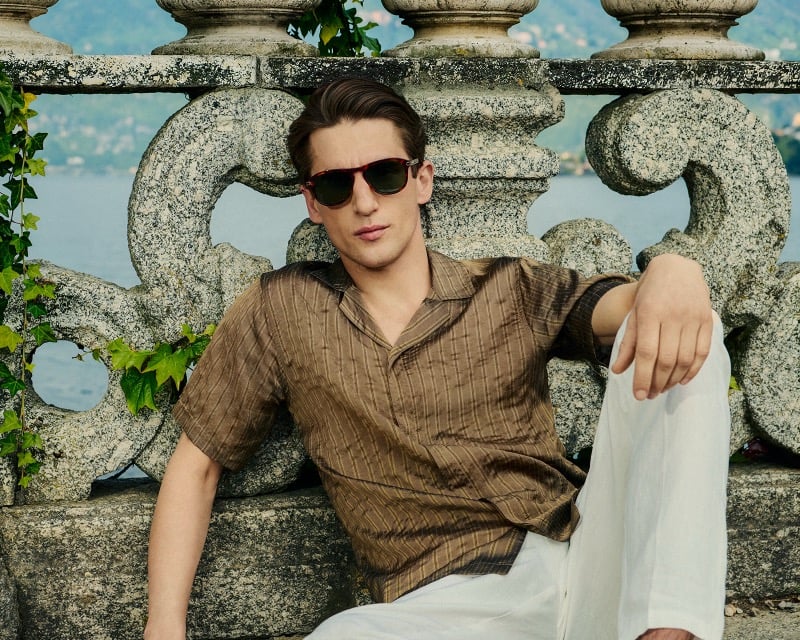Anatol Modzelewski sits against a carved stone balustrade wearing a brown striped camp-collar shirt and sunglasses