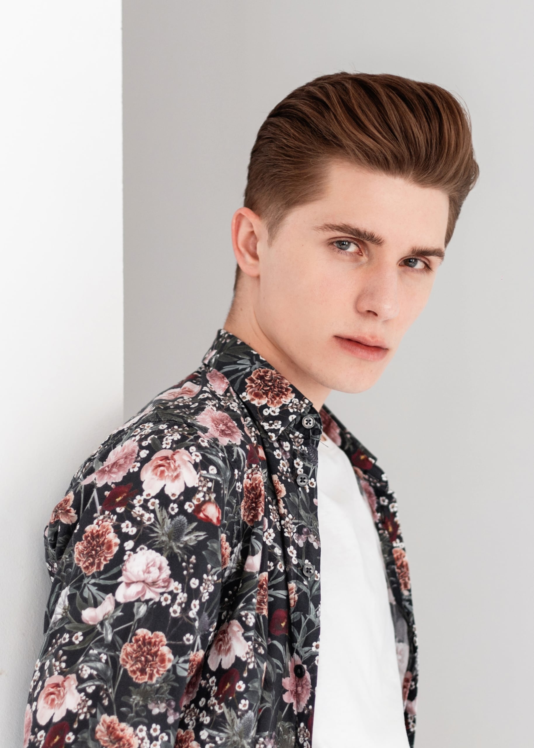 A young man in an oversized peony print shirt on a black ground leaning against a wall