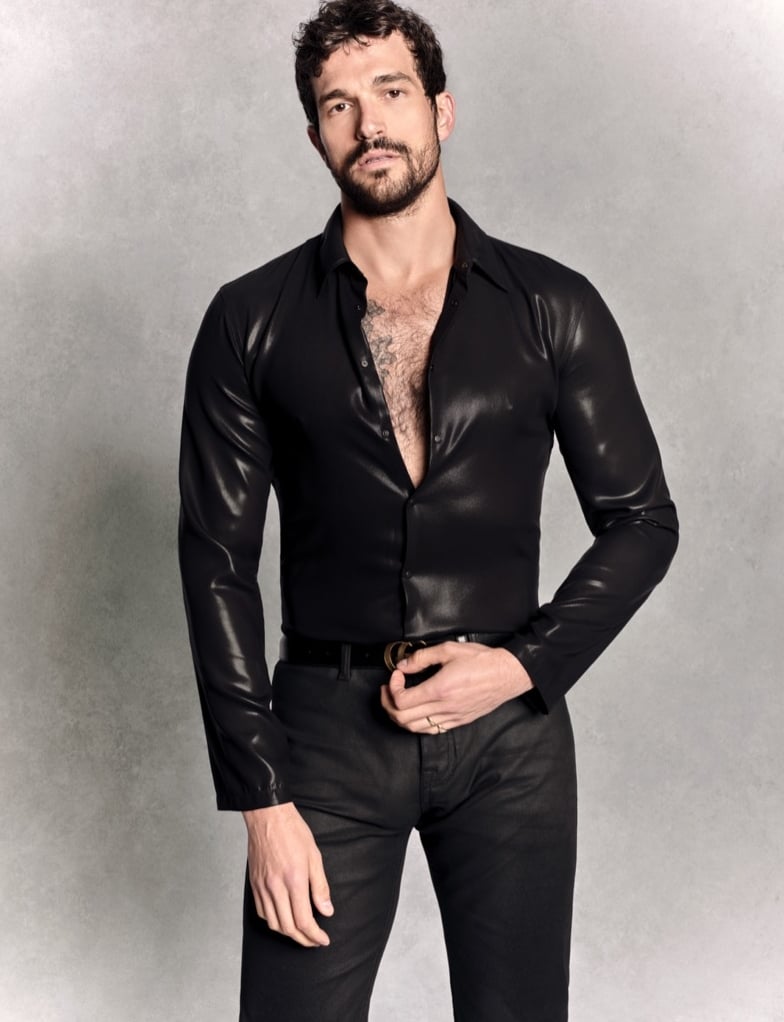 Paul Anthony Kelly in a satin black Gucci shirt with the top buttons undone, one hand on a logo belt buckle, tattoo visible on his chest