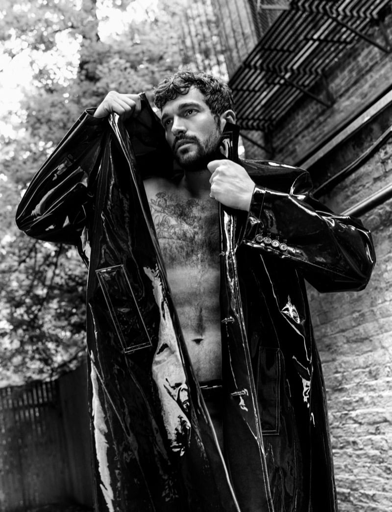 Paul Anthony Kelly in a patent leather Saint Laurent trench coat worn open over a bare, tattooed chest, photographed from a low angle outdoors