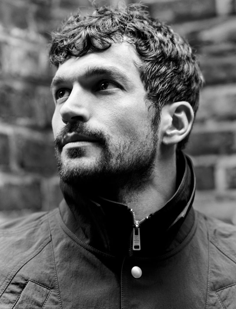 Close-up of Paul Anthony Kelly in a narrow brick alley wearing a double-breasted trench coat with a cape-like shoulder collar 