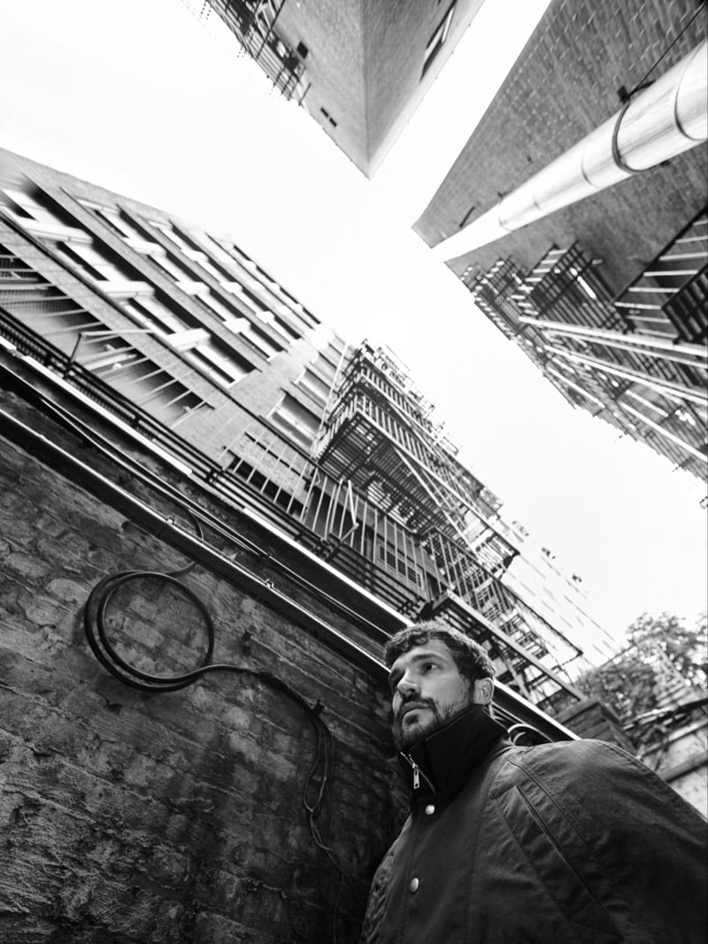 Paul Anthony Kelly stands in a brick alley looking upward, photographed from a low angle amid fire escapes and towering buildings