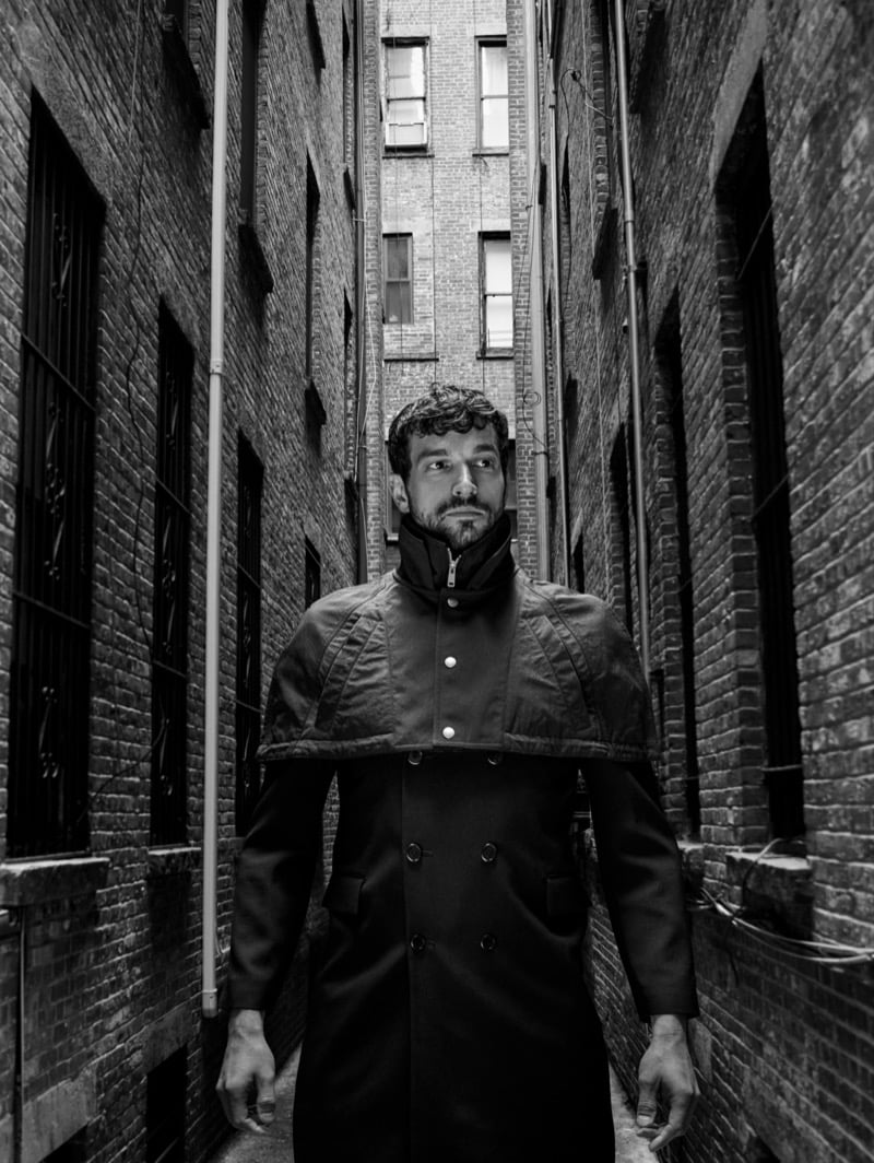Paul Anthony Kelly stands in a narrow brick alley wearing a double-breasted trench coat with a cape-like shoulder collar 