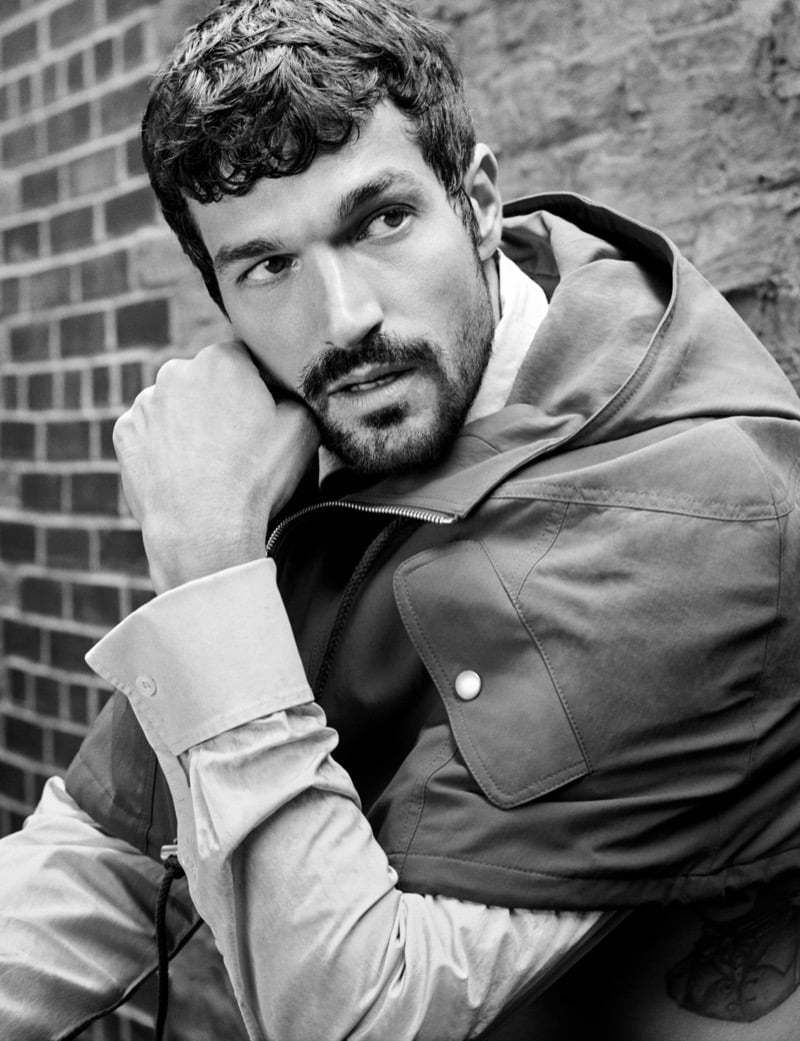 Paul Anthony Kelly rests his cheek on his fist in profile, wearing a utility jacket with a zip collar against a brick wall
