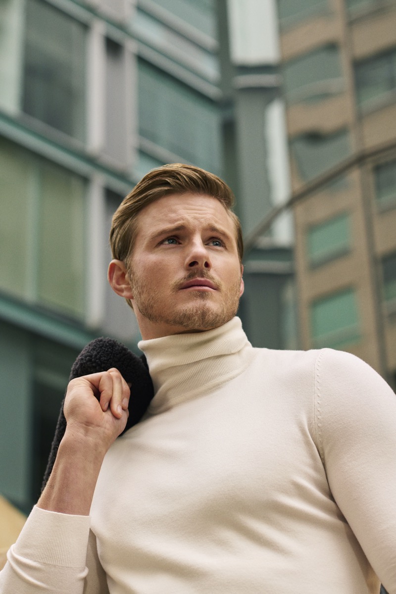 Alexander Ludwig in a cream turtleneck, a shearling-collar jacket slung over one shoulder, photographed against glass office buildings