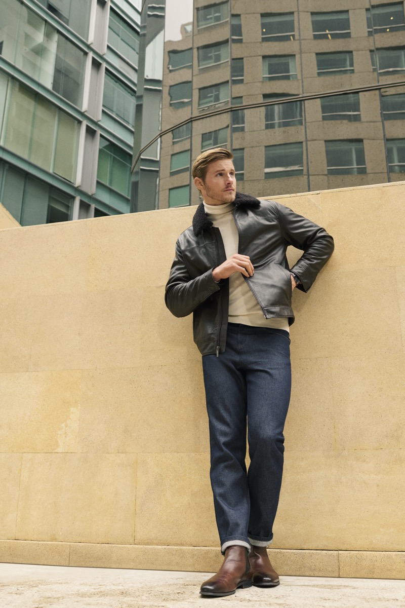 Alexander Ludwig in a shearling-collar leather jacket, denim, and brown chelsea boots, leaning against a stone wall