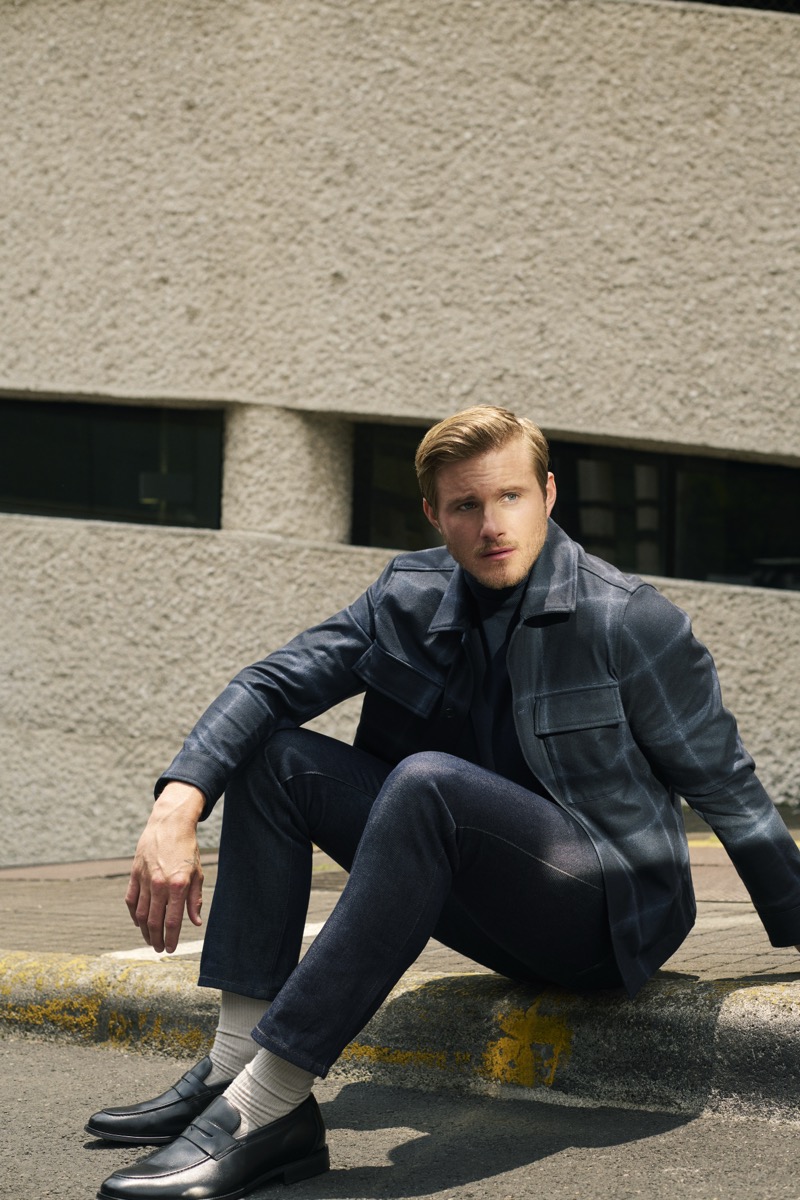 Alexander Ludwig seated on a curb in a checked wool overshirt, cropped trousers, and black loafers