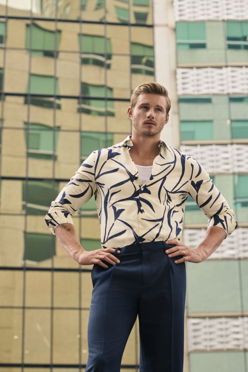 Alexander Ludwig in an open leaf-print shirt over a tank top and pleated trousers, hands on hips in front of a glass tower