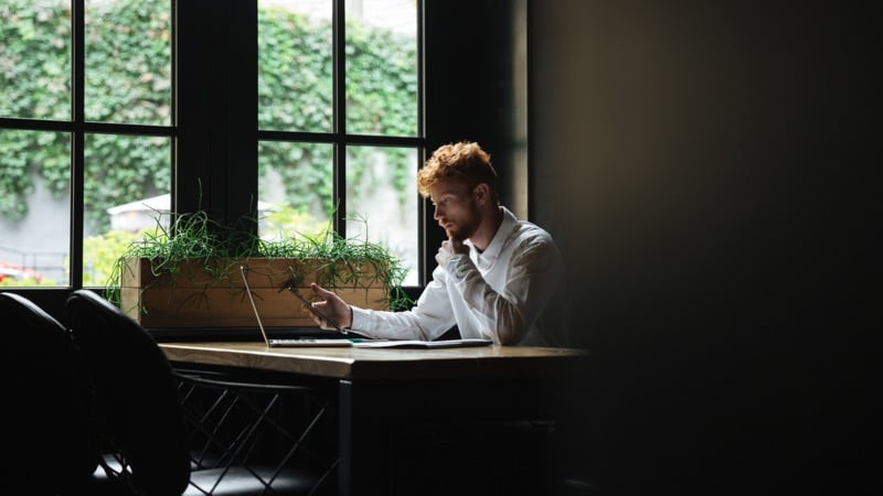 A bearded man in a white shirt checks his phone at a desk with an open laptop in a plant-filled office