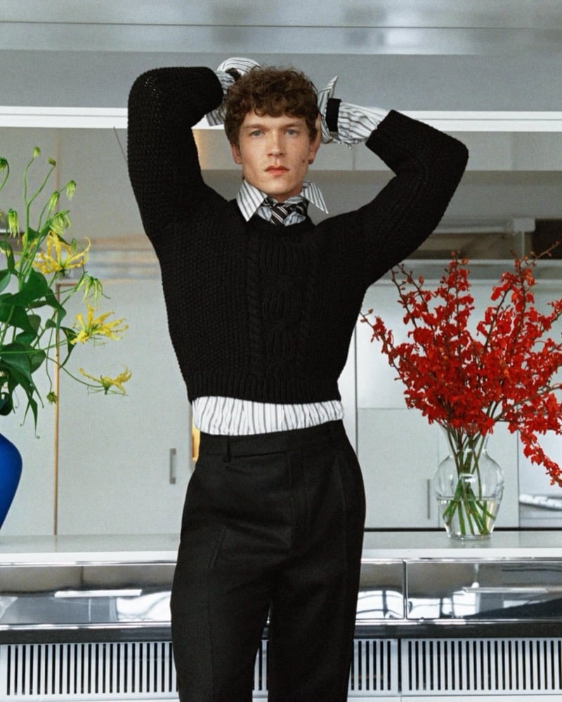 Connor Storrie wears a shrunken black cable-knit sweater, striped shirt and tie, and black trousers, arms raised, with flowers in the background for Saint Laurent's fall 2026 campaign.