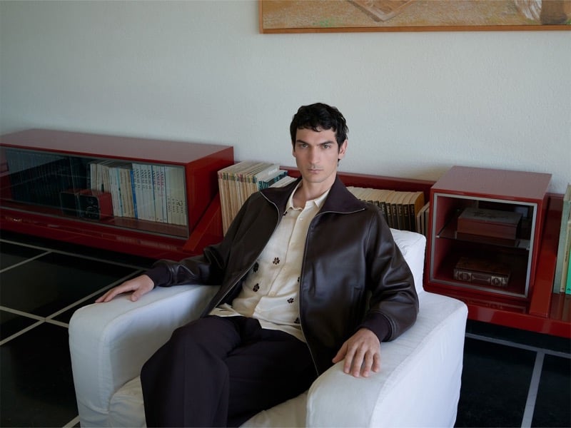 Luca Lemaire sits in a white armchair wearing a brown leather jacket over a cream printed shirt and dark trousers, red bookshelves behind him