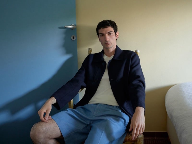 Luca Lemaire sits in a navy jacket over cream knitwear and blue shorts, one arm resting on a wicker chair